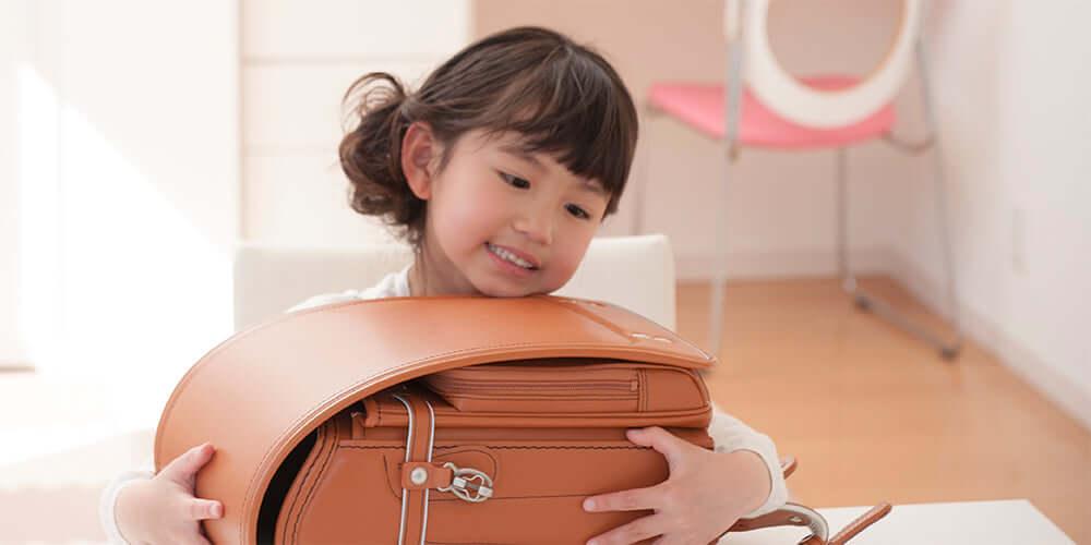 Smiling child holding a brown Japanese Randoseru backpack indoors