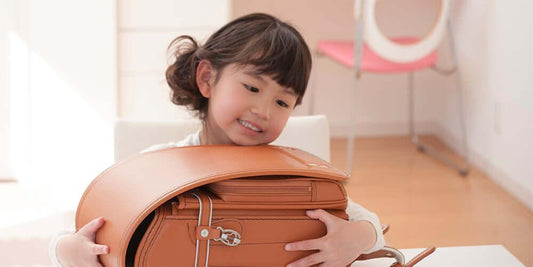 Smiling child holding a brown Japanese Randoseru backpack indoors