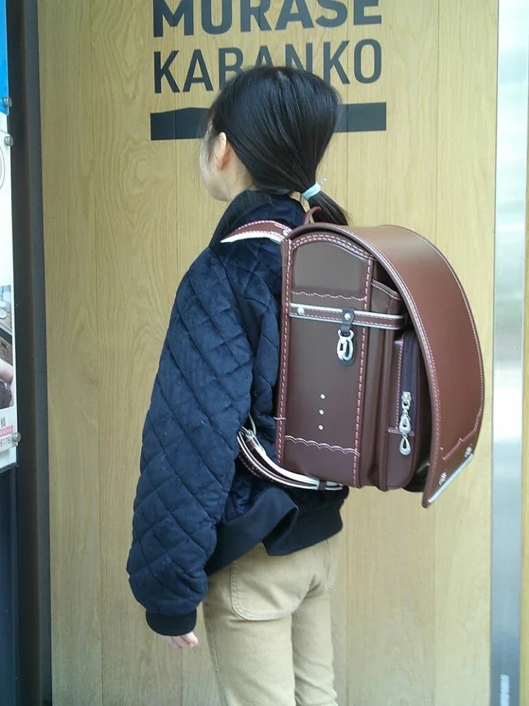 Child wearing brown Japanese randoseru backpack in front of wooden door